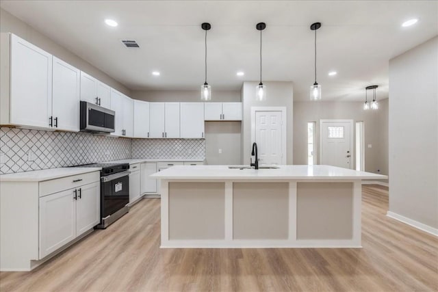 kitchen with white cabinetry, hanging light fixtures, stainless steel appliances, a kitchen island with sink, and light wood-type flooring