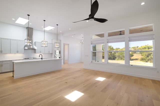 kitchen with light wood-type flooring, a skylight, wall chimney exhaust hood, pendant lighting, and an island with sink