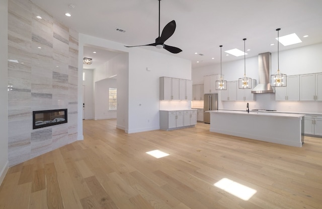 kitchen with high quality fridge, wall chimney exhaust hood, ceiling fan, hanging light fixtures, and an island with sink