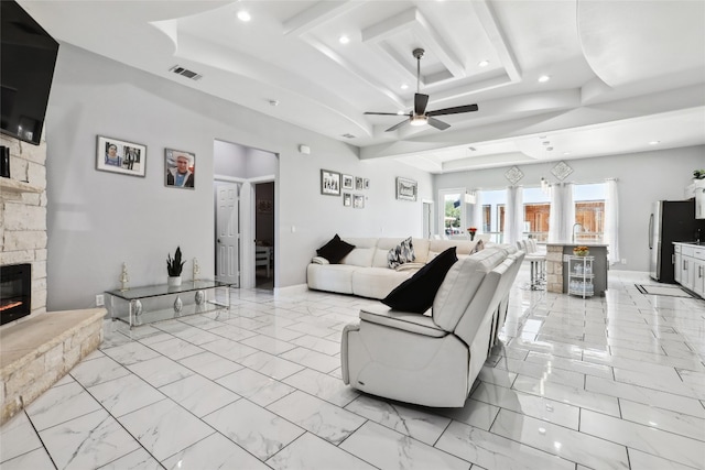 living room with a raised ceiling, a stone fireplace, and ceiling fan