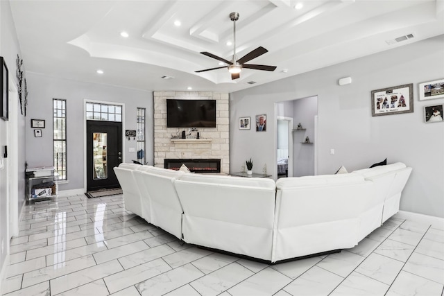 living room with a raised ceiling, a stone fireplace, and ceiling fan