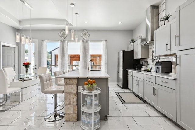 kitchen with hanging light fixtures, wall chimney range hood, a kitchen breakfast bar, gray cabinets, and a kitchen island with sink