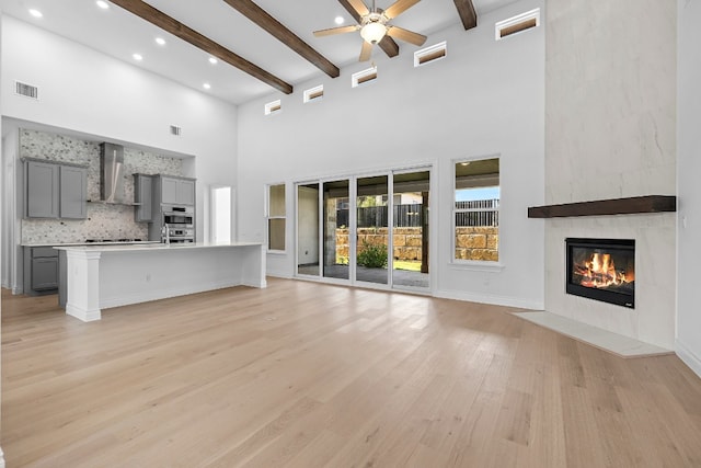 unfurnished living room with beam ceiling, ceiling fan, a large fireplace, a high ceiling, and light wood-type flooring