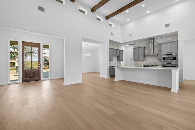 kitchen with a high ceiling, wall chimney exhaust hood, gray cabinets, light wood-type flooring, and an island with sink