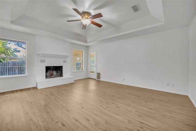 unfurnished living room featuring a tray ceiling, ceiling fan, light hardwood / wood-style floors, and a brick fireplace