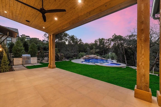 patio terrace at dusk featuring ceiling fan, an outdoor kitchen, a grill, and a lawn