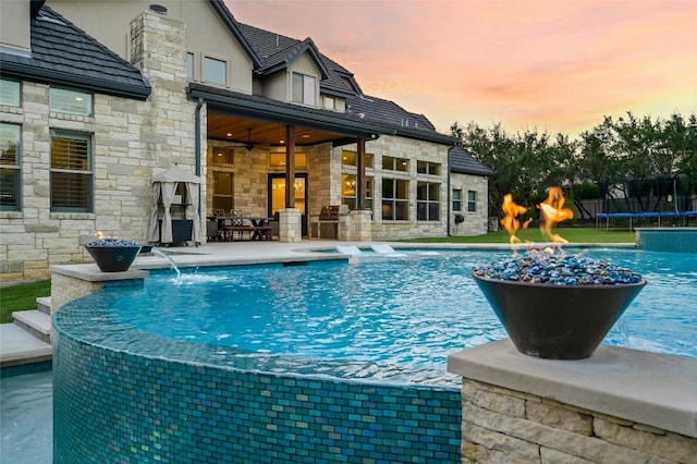 pool at dusk with a trampoline, pool water feature, ceiling fan, an in ground hot tub, and a patio area