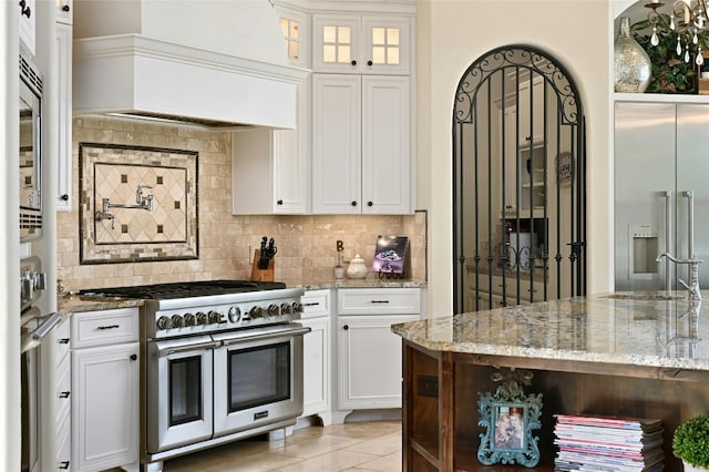 kitchen with tasteful backsplash, light stone counters, white cabinets, custom range hood, and high end appliances