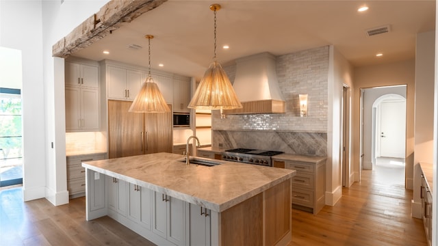 kitchen with light wood-type flooring, sink, custom exhaust hood, and an island with sink