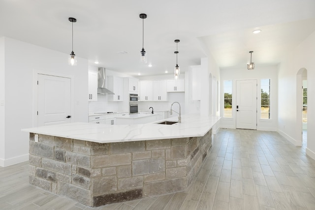 kitchen with light stone counters, wall chimney exhaust hood, sink, decorative light fixtures, and white cabinets