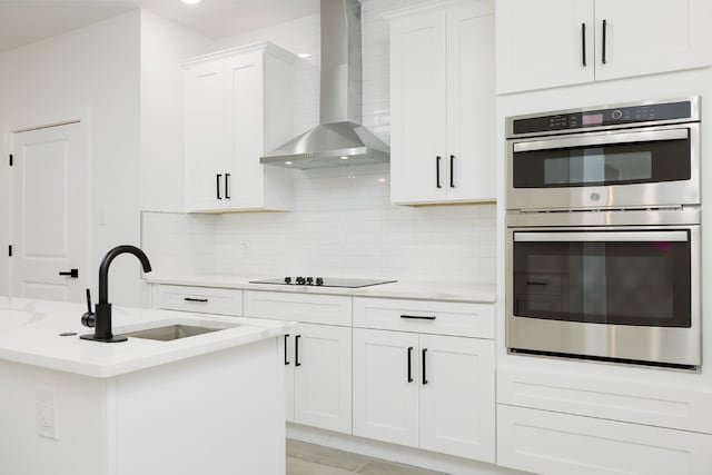 kitchen with white cabinetry, light stone countertops, sink, stainless steel double oven, and wall chimney range hood