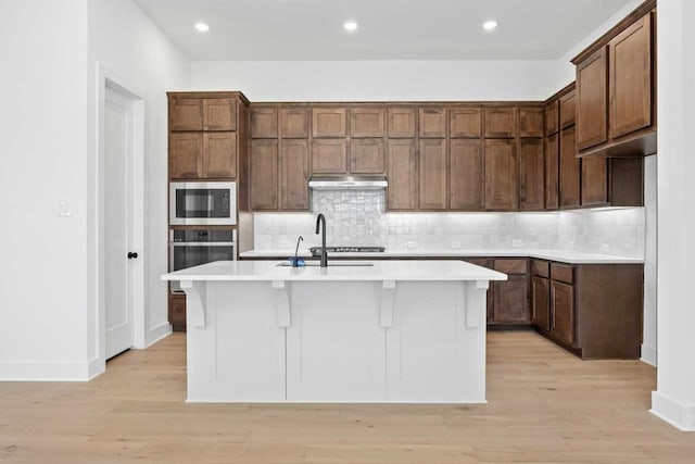 kitchen with built in microwave, oven, a center island with sink, and backsplash