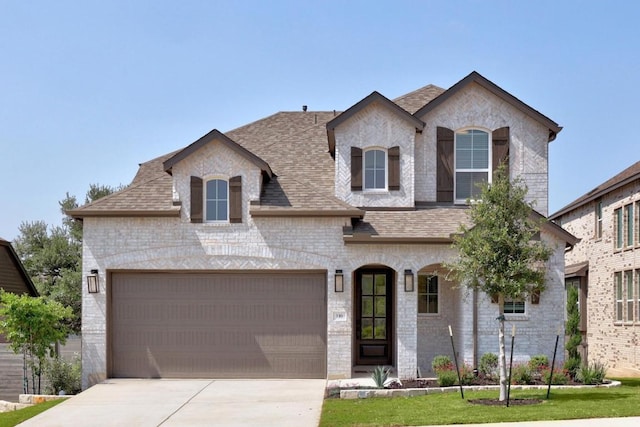 french country inspired facade featuring concrete driveway, brick siding, an attached garage, and a shingled roof