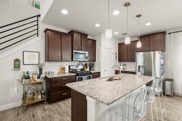 kitchen featuring an island with sink, light stone counters, hanging light fixtures, stainless steel appliances, and a sink