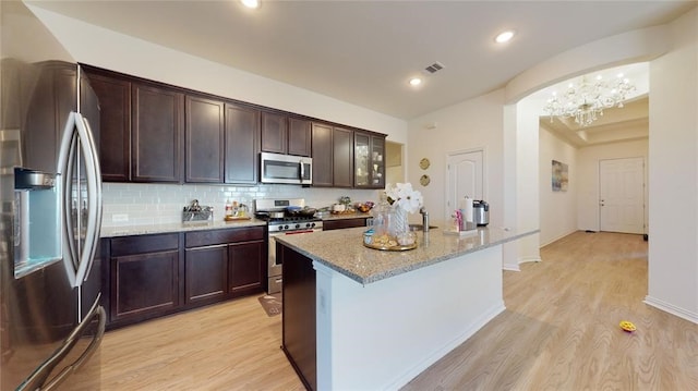 kitchen with light stone counters, light hardwood / wood-style flooring, stainless steel appliances, decorative backsplash, and a kitchen island with sink