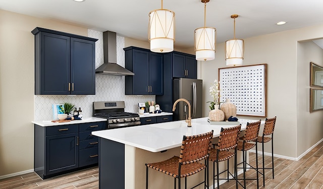 kitchen with hanging light fixtures, wall chimney range hood, an island with sink, a breakfast bar area, and appliances with stainless steel finishes