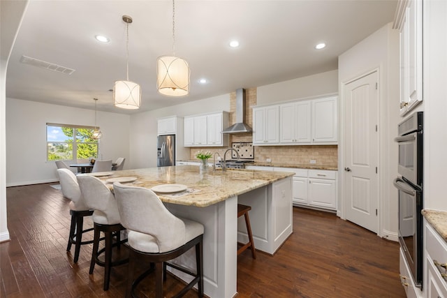 kitchen with appliances with stainless steel finishes, an island with sink, white cabinets, and wall chimney range hood