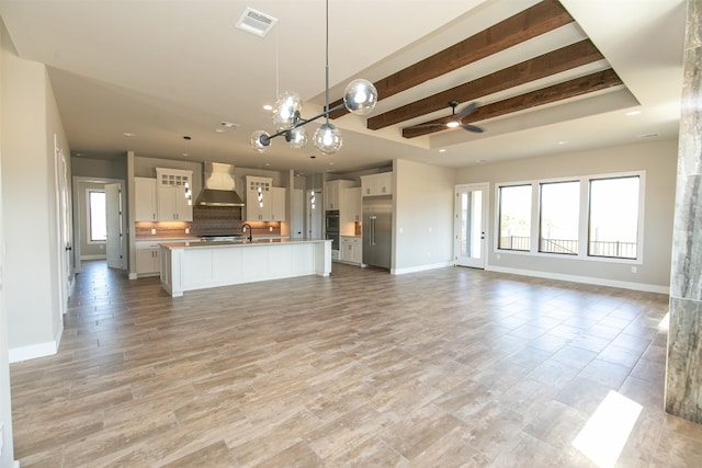 kitchen with beam ceiling, white cabinetry, wall chimney exhaust hood, pendant lighting, and a center island with sink