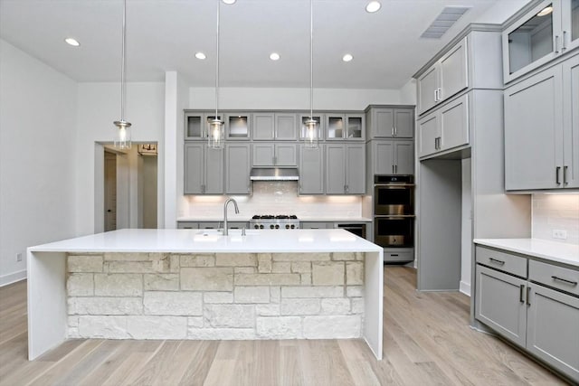 kitchen featuring decorative light fixtures, gray cabinets, stainless steel double oven, a kitchen island with sink, and decorative backsplash