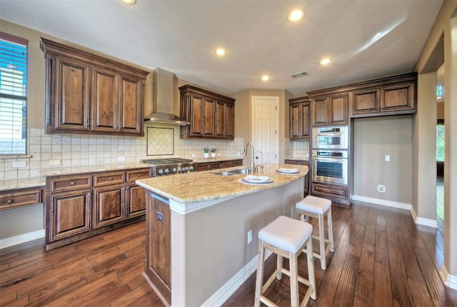 kitchen with light stone counters, a center island with sink, dark wood-type flooring, and wall chimney range hood