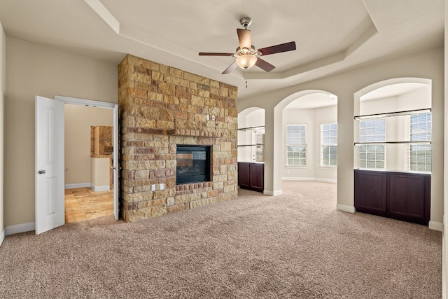 unfurnished living room with light colored carpet, a stone fireplace, ceiling fan, and a tray ceiling