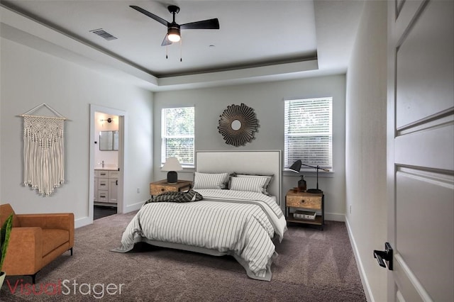 bedroom with ceiling fan, dark carpet, a tray ceiling, and ensuite bathroom