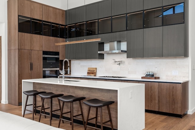 kitchen featuring sink, wall chimney range hood, black double oven, an island with sink, and light hardwood / wood-style floors