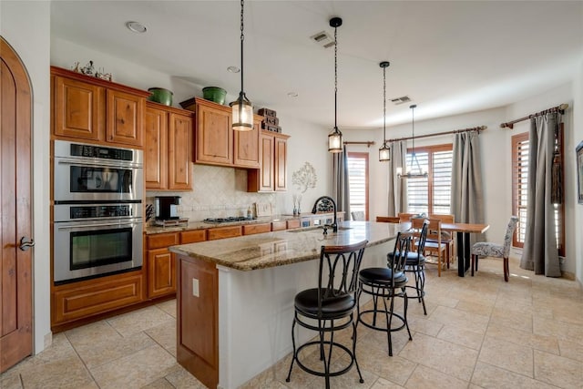 kitchen with an island with sink, sink, hanging light fixtures, light stone countertops, and stainless steel double oven