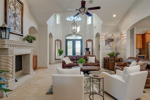 living room featuring french doors, light tile patterned flooring, a high end fireplace, ceiling fan with notable chandelier, and a high ceiling