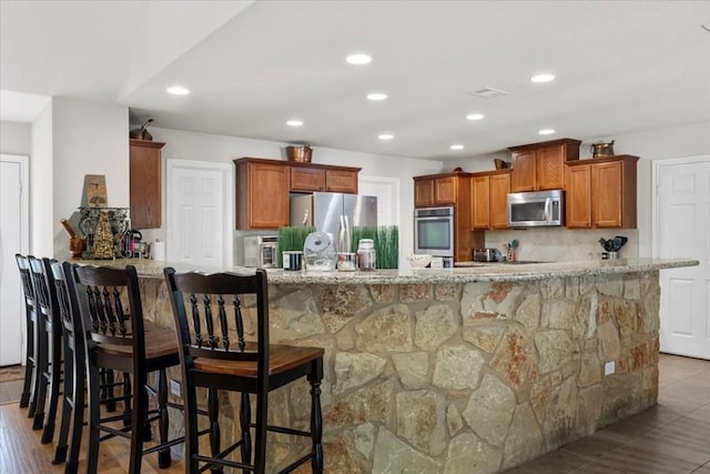 kitchen featuring a breakfast bar area, stainless steel appliances, brown cabinetry, light stone countertops, and a peninsula