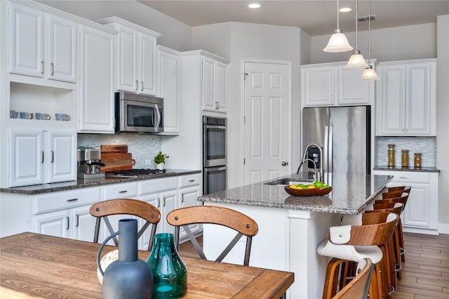 kitchen featuring stainless steel appliances, an island with sink, white cabinets, decorative light fixtures, and dark stone countertops