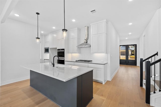 kitchen with light stone counters, wall chimney exhaust hood, stainless steel appliances, a center island with sink, and white cabinets