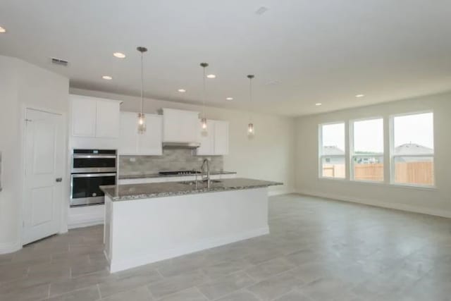 kitchen with pendant lighting, dark stone counters, white cabinets, a center island with sink, and double oven