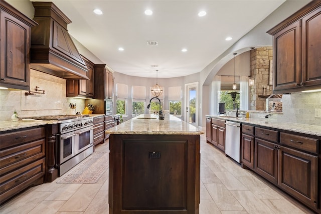 kitchen featuring an island with sink, light stone countertops, custom exhaust hood, stainless steel appliances, and a sink