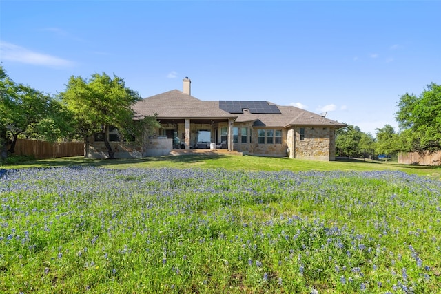 rear view of property featuring a chimney, solar panels, a lawn, fence, and stone siding