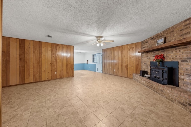 unfurnished living room with ceiling fan, a wood stove, a textured ceiling, and wooden walls