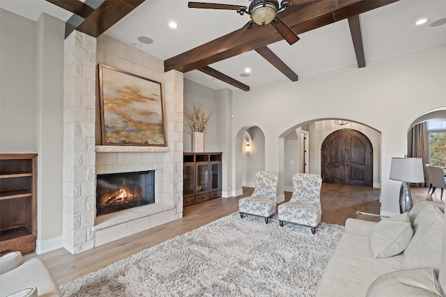 living room with beam ceiling, a stone fireplace, ceiling fan, and light wood-type flooring