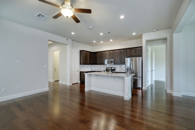 kitchen with light stone countertops, a kitchen island with sink, dark hardwood / wood-style flooring, decorative backsplash, and appliances with stainless steel finishes