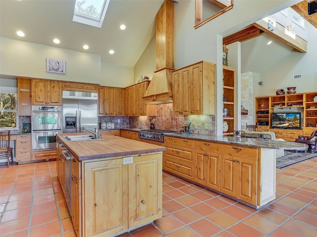 kitchen with a skylight, stainless steel appliances, high vaulted ceiling, a kitchen island with sink, and light tile patterned flooring