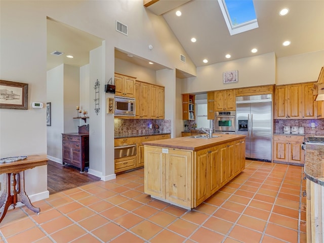 kitchen with a skylight, high vaulted ceiling, built in appliances, a center island with sink, and light tile patterned floors