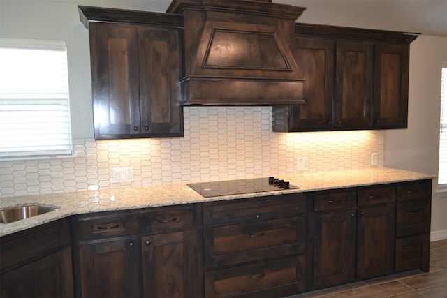 kitchen with dark brown cabinetry, light stone counters, premium range hood, decorative backsplash, and black stovetop