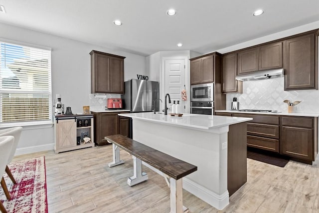 kitchen featuring decorative backsplash, light hardwood / wood-style floors, a kitchen island with sink, dark brown cabinets, and appliances with stainless steel finishes