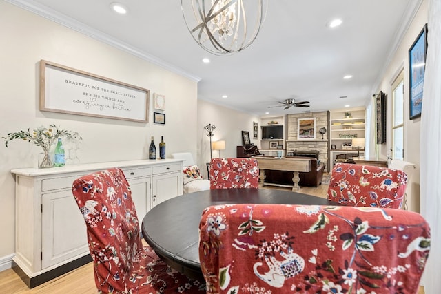 dining space with ceiling fan with notable chandelier, built in shelves, a stone fireplace, light hardwood / wood-style floors, and crown molding