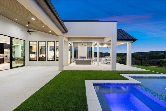 back house at dusk featuring a lawn, outdoor lounge area, ceiling fan, and a patio