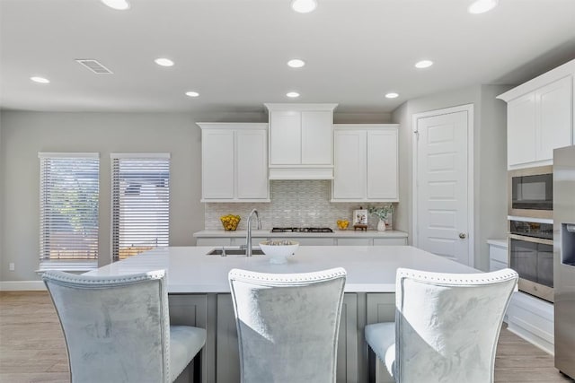 kitchen with white cabinetry, stainless steel appliances, backsplash, a kitchen island with sink, and light wood-type flooring