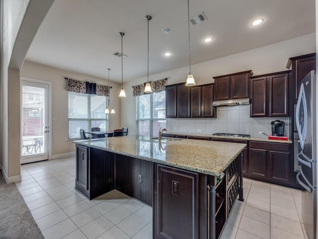 kitchen featuring decorative backsplash, appliances with stainless steel finishes, light stone counters, hanging light fixtures, and an island with sink