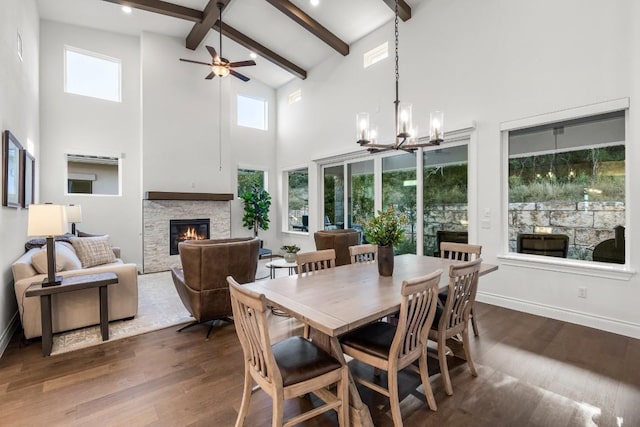 dining area featuring dark wood-type flooring, high vaulted ceiling, ceiling fan with notable chandelier, a fireplace, and beam ceiling