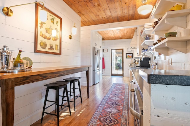 kitchen featuring wooden walls, stainless steel refrigerator, sink, wood ceiling, and light hardwood / wood-style flooring