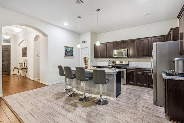 kitchen featuring light stone countertops, light hardwood / wood-style flooring, an island with sink, pendant lighting, and appliances with stainless steel finishes