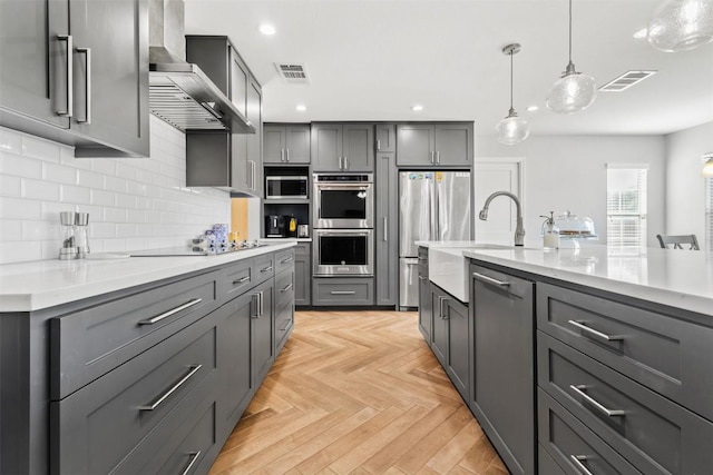 kitchen featuring wall chimney exhaust hood, pendant lighting, gray cabinets, light parquet flooring, and appliances with stainless steel finishes